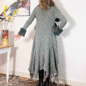 Könnte beinhalten: Ein langärmeliger, graumelierter Strickmantel mit dunkelgrauen Kunstfellbündchen und einem ausgestellten, asymmetrischen Saum. Das Kleidungsstück hat eine strukturierte Optik und wird mit schwarzen Stiefeln kombiniert, wobei ein Spitzenbesatz sichtbar ist.