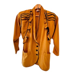 May include: Mustard-colored blazer with black trim and ruffles. The vintage jacket has a notched collar, three black buttons, and two front pockets. The blazer is hanging on a wooden hanger.