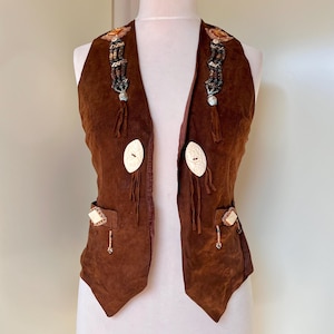 Peut inclure: Un gilet en daim marron avec des ornements décoratifs. Le gilet présente de grands boutons blanc cassé, des accents perlés et des détails de franges. Le gilet a deux poches et est présenté sur un mannequin.