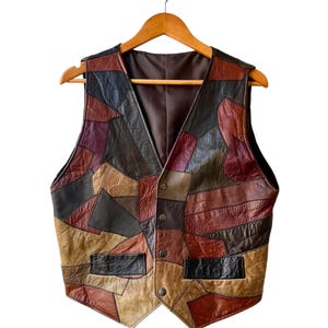 Op de afbeelding: Een vintage leren vest met een patchwork ontwerp. Het vest heeft een V-hals en twee voorzakken. Het kleurenpalet omvat tinten bruin, beige, bordeauxrood en zwart. Het hangt aan een houten kleerhanger.
