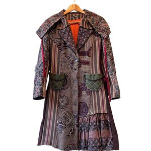May include: A long coat featuring a patchwork design with floral patterns, vertical stripes, and textured fabric panels. It includes two small olive green pockets, a wide collar, a bright orange interior lining, and a ruffled hem detail at the bottom.