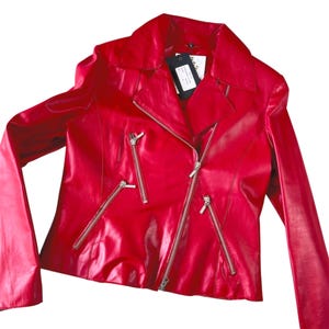 May include: A bright red jacket featuring a wide notched collar, long sleeves, and an asymmetrical front zipper closure. The jacket includes three silver-toned zippered pockets on the front and a black rectangular tag attached to the inner collar.