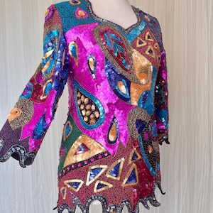 May include: A vibrant, sequined tunic in a kaleidoscope of colors, including fuchsia, teal, gold, and green. The long-sleeved top features an intricate pattern of paisley and geometric shapes, with a scalloped hemline and cuffs. The neckline is embellished with small beads.