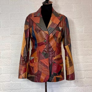 Puede incluir: Una chaqueta tipo blazer de manga larga confeccionada con parches de cuero multicolor en tonos marrón, rojo, naranja, azul y negro. La prenda presenta solapas de muesca, dos botones oscuros de cierre y dos bolsillos frontales de parche.
