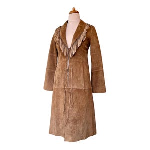 May include: A tan suede coat featuring long fringe detailing along the lapels and chest area. The coat has long sleeves, a knee-length hem, and a front closure system consisting of thin leather ties that fasten at the centre of the garment.