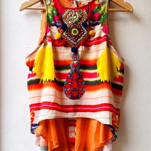 Puede incluir: Una blusa sin mangas con un diseño vibrante y colorido. La blusa presenta rayas horizontales en rojo, naranja, blanco y verde. Tiene un adorno de cuentas y borlas amarillas. La capa inferior es de tela naranja fluida.