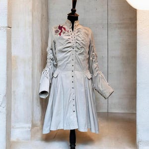 May include: A light beige button-down coat featuring a high collar, ruched detailing along the centre front, and long sleeves with adjustable drawstring ties. A small, detailed embroidery of a dark red bird is positioned on the upper right chest.