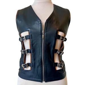 Peut inclure: Gilet en cuir noir avec un col en V et une fermeture éclair argentée. Le gilet présente des découpes avec des sangles noires et des boucles argentées. L'intérieur est doublé de couleur beige. Le gilet est présenté sur un mannequin.