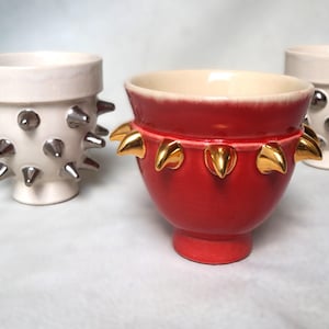 May include: Three ceramic vessels. Two are white with silver spikes, and one is red with gold leaf accents. The vessels have a unique, decorative design, suitable for home decor or as artistic pieces.