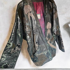 May include: A dark green and black patchwork jacket featuring various textured leather panels, embroidered rose motifs, and a deep burgundy interior lining. The garment has long sleeves, an open front design, and a complex surface pattern.