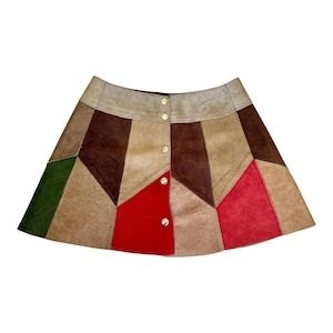 Może przedstawiać: Mini spódnica w stylu patchwork w różnych kolorach, w tym brązowym, beżowym, czerwonym i zielonym. Spódnica wykonana jest z zamszu lub podobnego materiału i posiada zapinany na guziki przód ze złotymi guzikami. Spódnica ma geometryczny wzór.