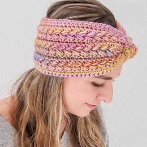 May include: A hand-crocheted headband in shades of pink, yellow, and orange. The headband has a textured, braided pattern and is worn across the forehead. The soft colours and intricate design suggest a cosy, stylish accessory, perfect for cooler weather.