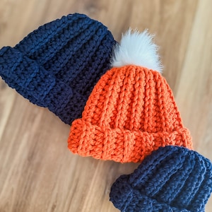 May include: Three handmade crochet beanies in navy blue and orange. The orange beanie has a white faux fur pom-pom on top. The beanies are made with a thick yarn and have a ribbed texture.