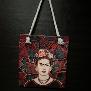 Puede incluir: Un bolso tote con un retrato de Frida Kahlo. El bolso tiene un fondo estampado en rojo y verde. El retrato muestra a Frida con una corona de flores y un collar. El bolso tiene asas de cuerda blanca.