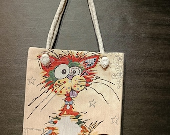 May include: A beige tote bag with a whimsical cat design. The cat has a colorful, expressive face with red, green, and yellow fur. The bag features a white rope handle and is decorated with stars and floral accents.