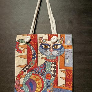 May include: A patterned tote bag with a cat design. The bag has a white rope handle and a rectangular shape. The cat illustration is made up of various patterns and colours, including blue, red, orange, and green.