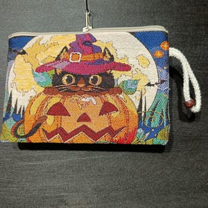 May include: A Halloween-themed tapestry wallet featuring a black cat wearing a witch hat, sitting inside a carved pumpkin. The background includes a full moon and colorful foliage. The wallet has a zipper closure and a white cord.