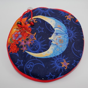 May include: Round, celestial-themed fabric item with a navy blue background, featuring a crescent moon face, stars, and suns. The item has a red border and a floral and star design on the left side. A red drawstring is attached.