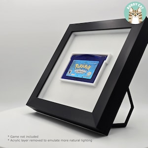 Può includere: Cartuccia di gioco Pokémon Versione Zaffiro incorniciata. La cartuccia blu è inserita in una cornice bianca, all'interno di una cornice nera. La cornice è appoggiata su una superficie bianca. Il titolo del gioco è visibile sulla cartuccia.