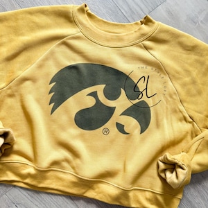 May include: Mustard yellow sweatshirt with a dark green graphic of a hawk. The sweatshirt has a crew neck and long sleeves. The text "THE STYLE LOFT" is printed on the right side of the hawk graphic.