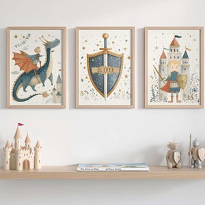 May include: Three framed watercolor prints with a fairytale theme. One shows a dragon with a rider, another a shield and sword with the name "Oliver", and the third a knight and castle. A wooden shelf below holds a toy castle and other decorative items.