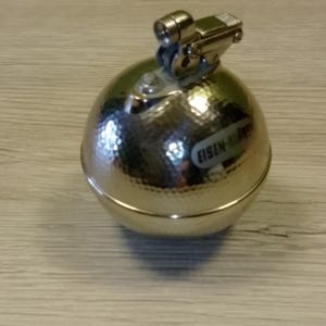 May include: A silver-colored, spherical lighter with a textured surface. The lighter has a hinged top with a small, rectangular component and a cylindrical nozzle. The text "EISEN-KRAFT" is visible on the side of the lighter.