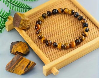May include: A bracelet made of polished tiger's eye beads, displayed on a small wooden tray. The beads are a mix of golden brown and dark brown. Several raw tiger's eye stones are also present.
