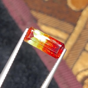 May include: A rectangular gemstone held by tweezers. The gem features a gradient of colors, transitioning from red at the ends to yellow and green in the center. The gem is faceted, reflecting light.