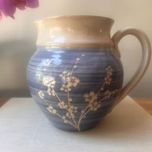 May include: A ceramic pitcher with a blue body and a cream-colored rim and handle. The blue section features a floral design in white. The pitcher is sitting on a white surface, with a blurred pink flower in the background.