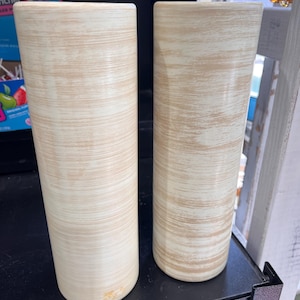 Op de afbeelding: Twee cilindrische vazen met een crèmekleurige basis en een textuur, lichtbruin, horizontaal streepjespatroon. De vazen zijn hoog en hebben een eenvoudig, modern design. Ze worden op een zwart oppervlak weergegeven.