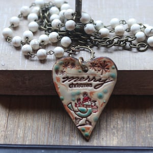 May include: A necklace featuring a heart-shaped pendant with a cream-colored glaze, the text merry christmas, and a graphic of a bird wearing a hat and scarf. The pendant hangs from a chain of white stone beads and dark bronze-toned metal links.