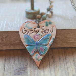 May include: A heart-shaped pendant featuring a blue butterfly and the text "Gypsy Soul" in raised lettering. The pendant has a light tan base with pink and teal accents and is attached to a beaded chain with small, round, multi-colored beads.