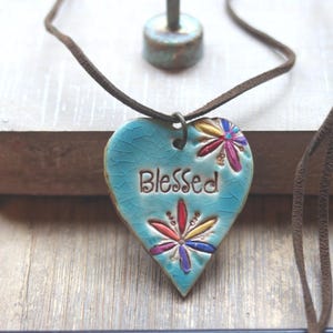 May include: A heart-shaped ceramic pendant in a light blue crackle glaze finish, featuring the word Blessed stamped in the center. The pendant is decorated with two colorful floral motifs in red, yellow, and purple, and hangs from a brown cord.