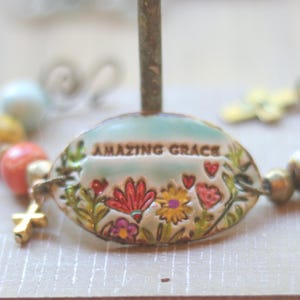 May include: A bracelet featuring a central oval ceramic charm with the text "AMAZING GRACE" above a colorful floral design. The bracelet includes multi-colored ceramic beads in shades of blue, yellow, and red, along with a small gold-toned cross charm.