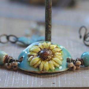 May include: A bracelet featuring a central ceramic sunflower charm in yellow and turquoise, flanked by alternating turquoise and yellow ceramic beads, small brown accent beads, and a dark metal wire clasp mechanism.