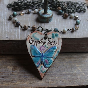 May include: A heart-shaped pendant necklace featuring a blue butterfly and the text "Gypsy Soul" in black lettering. The pendant is made of a cream-colored material with a crackled finish and is attached to a dark metal chain with multi-colored beads.