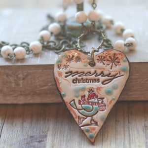 May include: A heart-shaped pendant necklace featuring the embossed text merry christmas, a bird illustration, and snowflake accents. The pendant is cream-colored with blue and red details, suspended from a bronze chain with white beads.