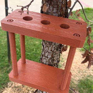 Puede incluir: Un columpio de madera marrón rojizo con un aspecto de veta de madera texturizada. El columpio tiene un asiento rectangular y una pieza superior con tres agujeros circulares. Cuatro soportes cilíndricos conectan el asiento a la pieza superior. El columpio está colgado de un árbol.