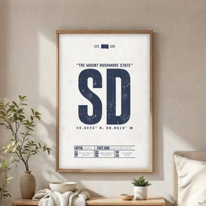 Könnte beinhalten: Ein gerahmter Druck mit den Buchstaben "SD" in Marineblau, mit dem Text "The Mount Rushmore State" darüber. Der Druck enthält Koordinaten und Fakten, alles vor einem strukturierten, cremefarbenen Hintergrund. Der Rahmen ist helles Holz.