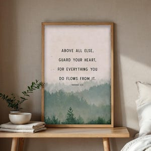 May include: A framed print with the text "ABOVE ALL ELSE, GUARD YOUR HEART, FOR EVERYTHING YOU DO FLOWS FROM IT. PROVERBS 4:23" against a backdrop of a misty forest. The frame is light brown, and the print is displayed on a wooden table.
