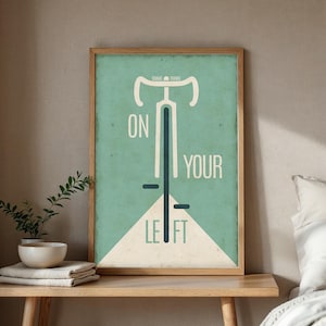 Könnte beinhalten: Ein gerahmter Kunstdruck mit einem Fahrrad-Design und den Worten "ON YOUR LEFT" in Weiß. Der Hintergrund ist helltürkis mit einem cremefarbenen Dreieck am unteren Rand. Der Rahmen ist aus Holz.
