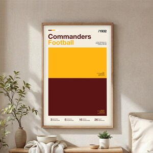 May include: A framed poster with a minimalist design. The poster features the text "Commanders Football" in yellow and white, with a gold and burgundy colour block design. The poster also includes the year "/1932" and other text.
