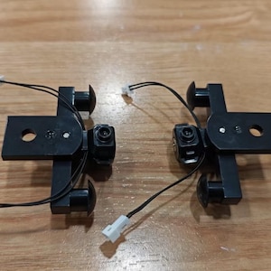 Puede incluir: Dos soportes de cámara negros con cables y conectores blancos. Cada soporte tiene una base redonda y una placa rectangular con un agujero en el centro.