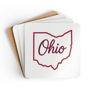 Puede incluir: Posavasos cuadrados blancos con un contorno burdeos del estado de Ohio y la palabra "Ohio" en cursiva. Los posavasos están apilados sobre un posavasos de corcho.