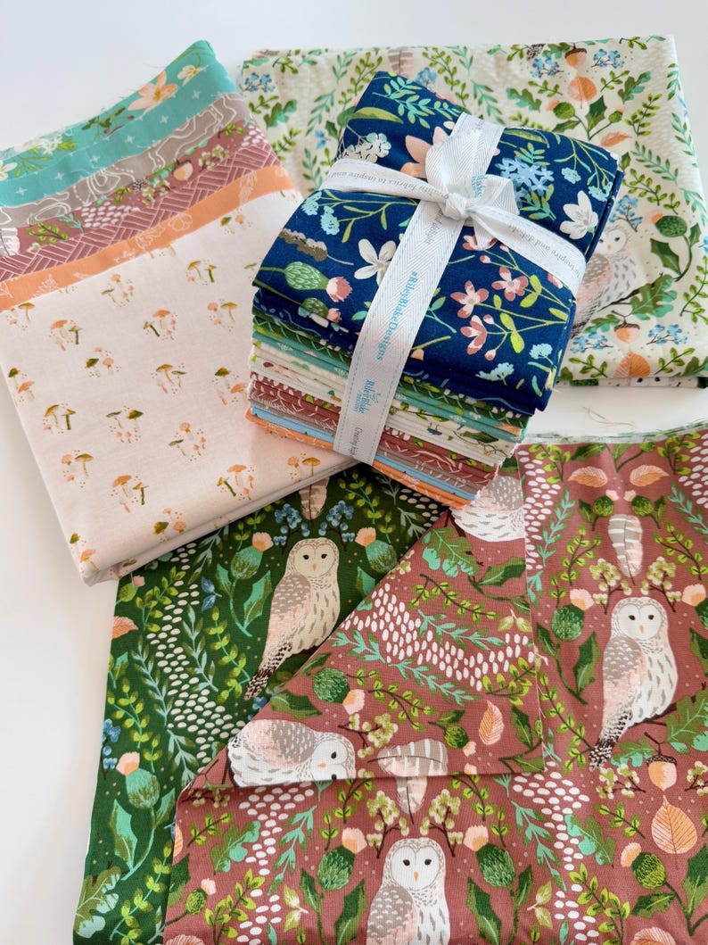 May include: A collection of patterned fabric bundles. The fabrics feature floral, mushroom, and owl designs in various colors, including blue, green, and peach. A white ribbon wraps around a stack of the fabric bundles.