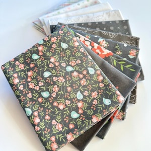 May include: A stack of fabric squares in various patterns and colors. The top square features a dark gray background with a floral and bird design in pink and teal. Other fabrics include floral, geometric, and solid patterns.