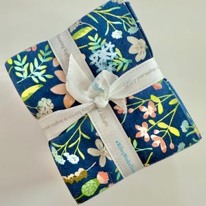 May include: A bundle of floral fabric tied with a white ribbon. The fabric has a navy blue background with a pattern of flowers in shades of pink, white, and yellow. The ribbon has text that reads "Creating high quality fabrics to inspire and delight."