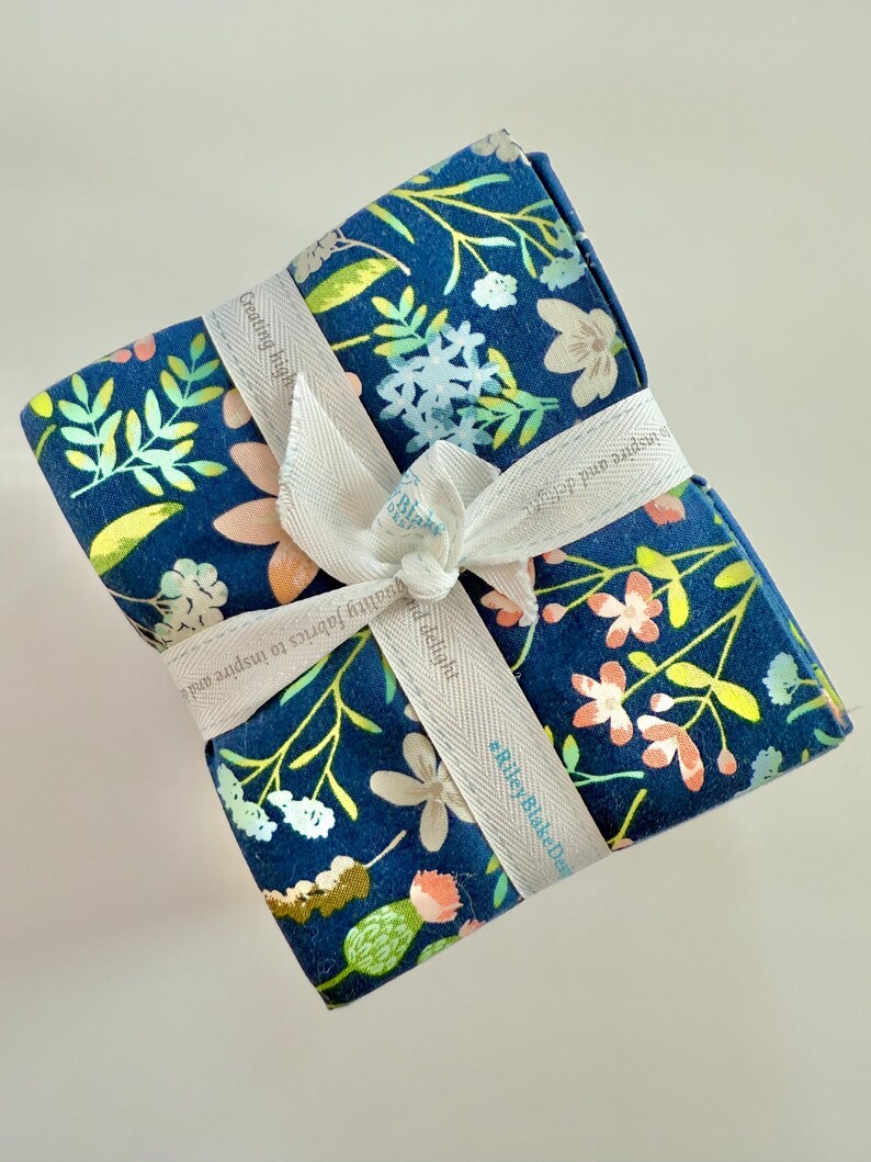 May include: A bundle of floral fabric tied with a white ribbon. The fabric has a navy blue background with a pattern of flowers in shades of pink, white, and yellow. The ribbon has text that reads "Creating high quality fabrics to inspire and delight."
