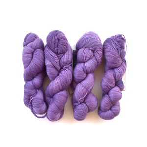 May include: Four skeins of lavender-colored yarn are arranged on a white background. The yarn is tightly wound into a twisted shape, with a soft, textured appearance. The color is a muted purple, with subtle variations in tone.