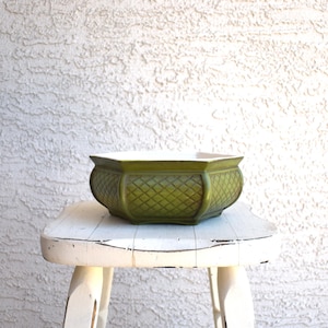 May include: A green ceramic planter with a diamond pattern sits on a white, distressed stool. The planter has a wide, octagonal shape with a white interior. The exterior is a muted green color, and the stool has a worn, vintage appearance.